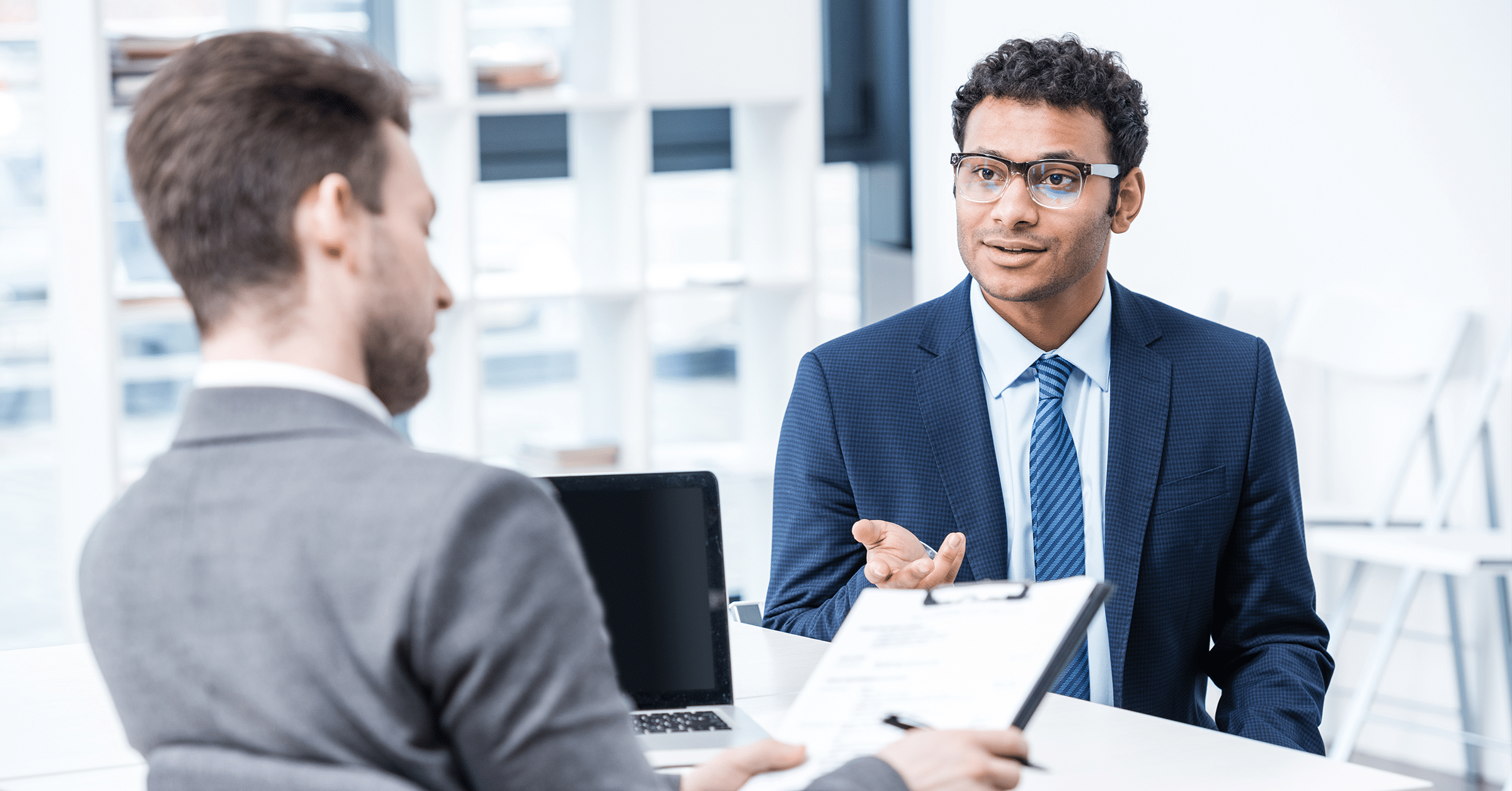 a man sits at his desk while holding a clipboard while an employee speaks to him